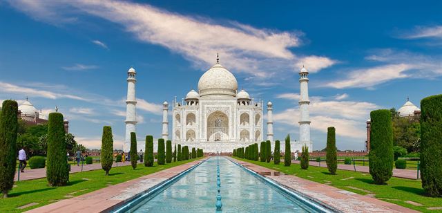 Wide view of the Taj Mahal beyond its reflecting pool and cypress-lined gardens under blue sky