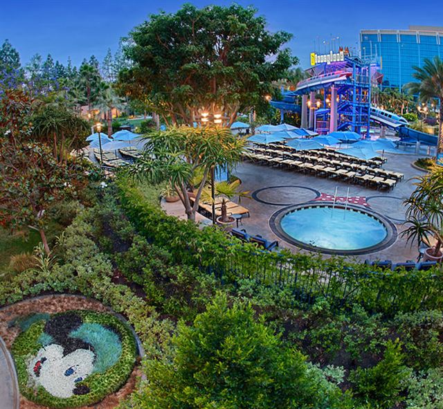 Disneyland Hotel pool area with Disneyland-sign waterslide tower, palms and a Mickey topiary at dusk
