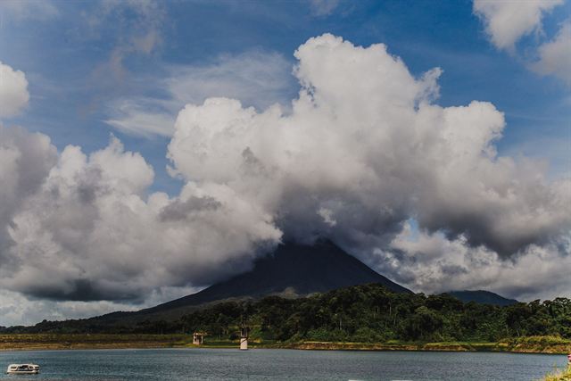 Arenal Volcano's cone shrouded in towering white clouds above Lake Arenal and forested shore