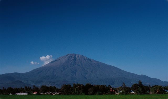 Volcanic cone of Mount Meru rising above trees and farmland under clear blue sky near Arusha