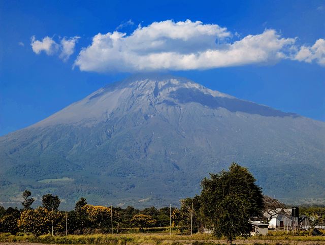 Mount Meru with a cloud over its summit above green trees and farmland near Arusha, Tanzania