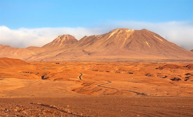 Atacama Desert plain with a winding track leading to brown volcanic mountains under blue sky