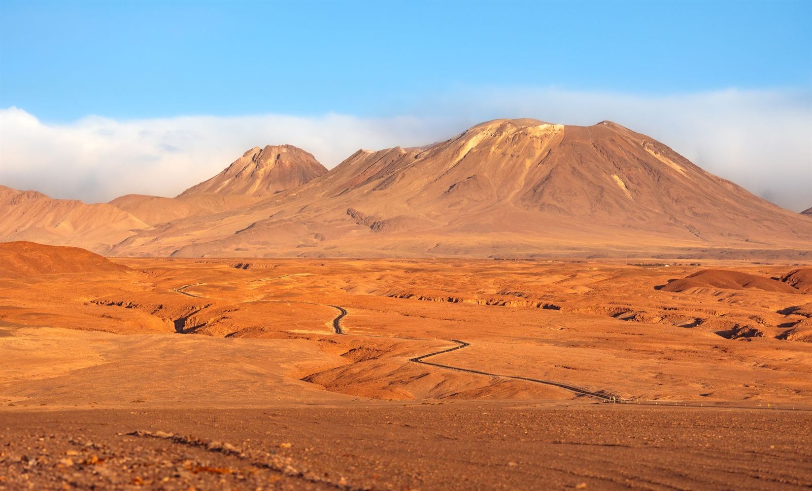Atacama Desert plain with a winding track leading to brown volcanic mountains under blue sky