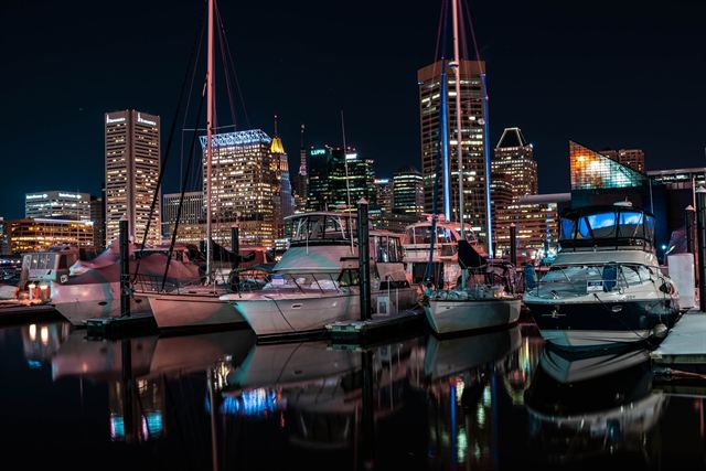 Sailboats and yachts moored in a Baltimore Inner Harbor marina at night with city towers behind