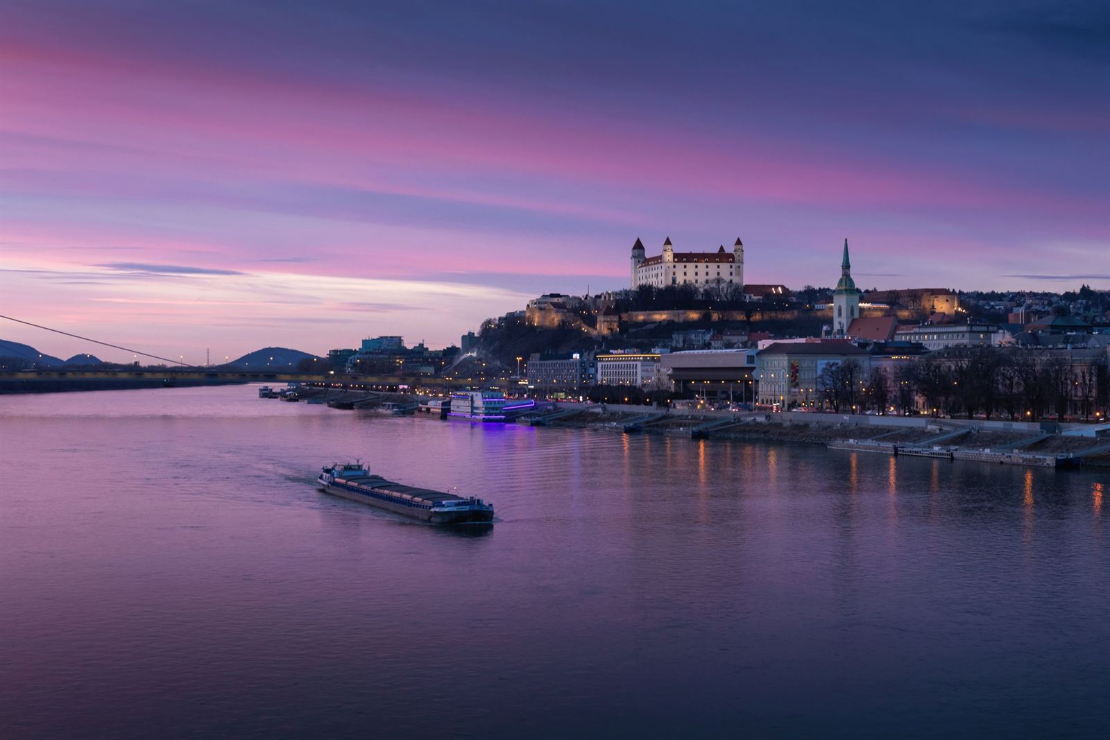 Bratislava Castle and St Martin's Cathedral lit above the Danube at purple dusk with a river barge