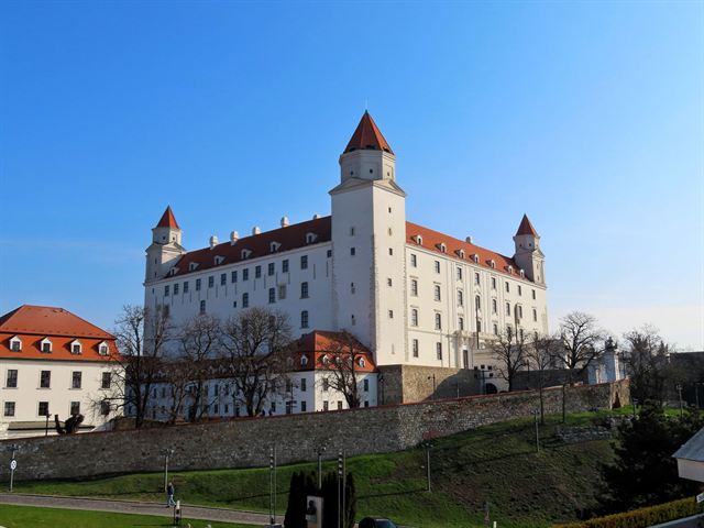 White four-towered Bratislava Castle with red roofs on its hill under blue sky