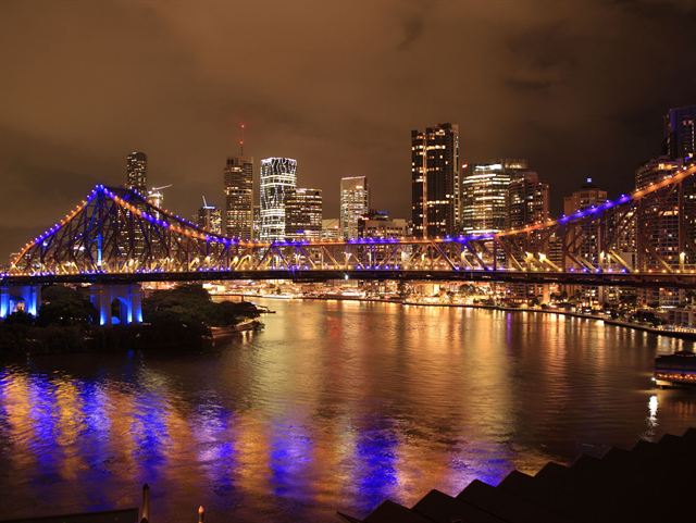 Story Bridge lit in blue and orange at night with Brisbane skyline reflected in the river