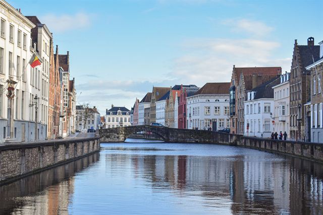 Calm Bruges canal reflecting brick gabled houses and a stone bridge under blue sky
