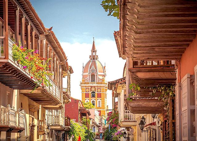 Colonial street with flower-draped wooden balconies leading to a cathedral bell tower in Cartagena