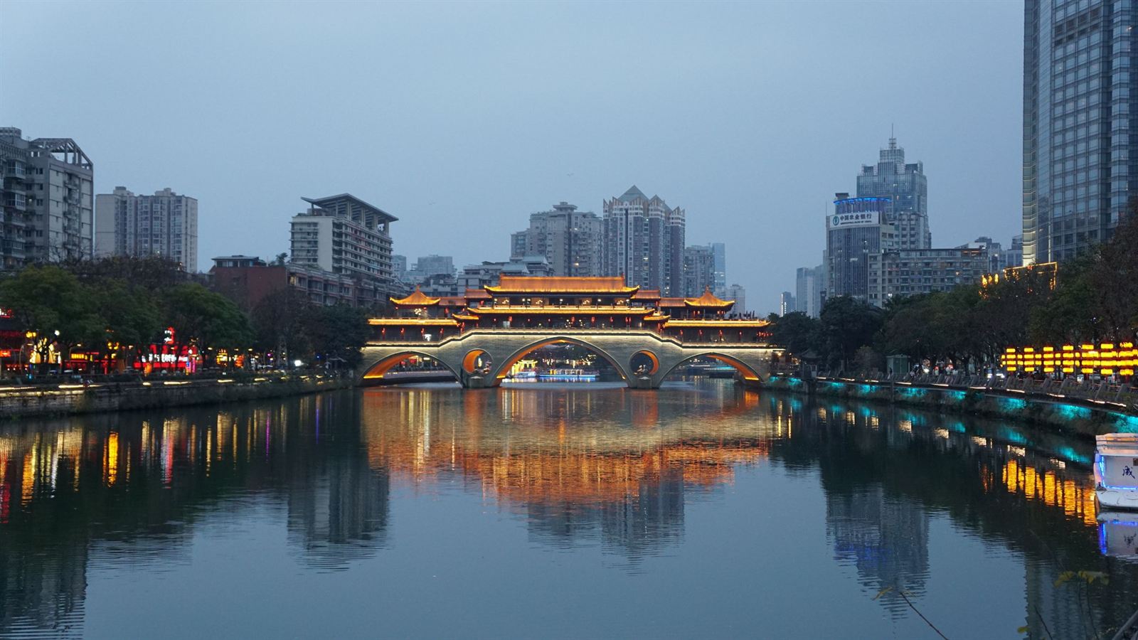 Anshun Bridge, a lit wooden covered bridge over the Jin River in Chengdu, with city towers at dusk