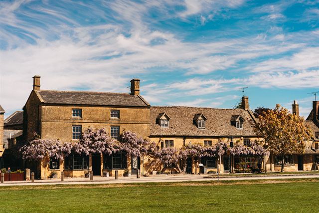 Cotswold stone inn draped in purple wisteria beside a village green in spring