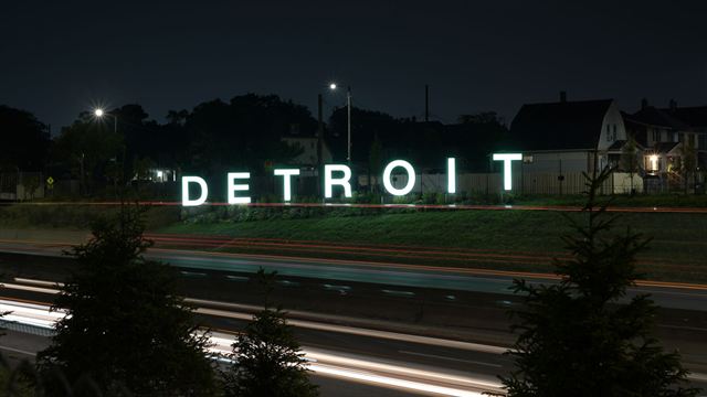 Illuminated DETROIT letter sign on a grassy slope at night beside a freeway with light trails
