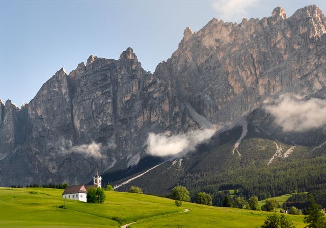 Small white church in a green meadow beneath towering grey Dolomites peaks