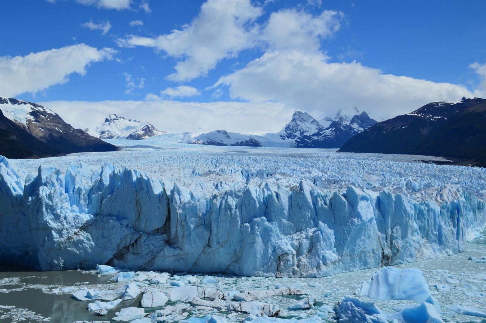 Jagged blue ice wall of Perito Moreno Glacier flowing from snowy Andes peaks under a bright sky