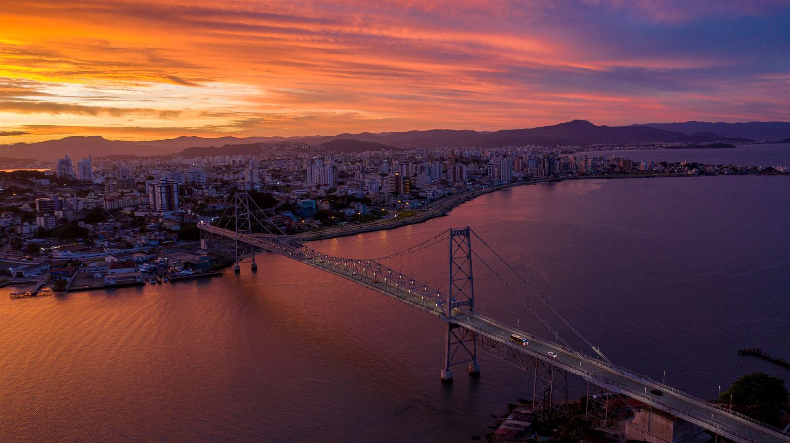Hercilio Luz suspension bridge spanning the bay to Florianopolis island under an orange sunset sky