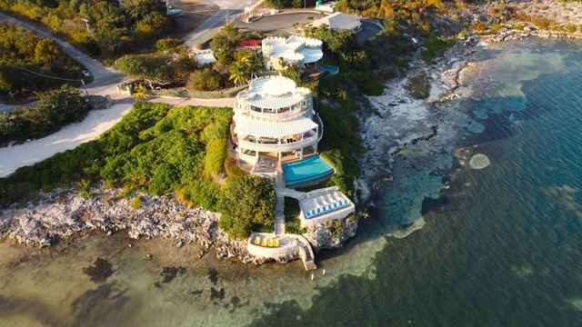 Aerial view of a round white seafront villa with pools on rocky ironshore and clear shallows, Grand Cayman