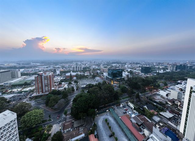 Aerial view over Guatemala City's high-rises, streets and treetops under a pastel sunset sky