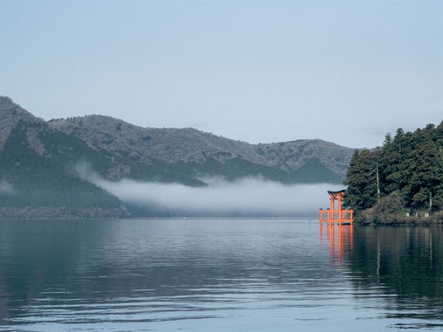 Red torii gate of Hakone Shrine standing in misty Lake Ashi below forested hills