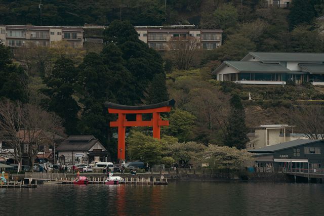 Red Hakone Shrine torii gate on the Lake Ashi shore beneath wooded hillside and hotels