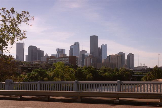 Downtown Houston skyline of high-rise towers seen over trees from a bridge railing in daylight
