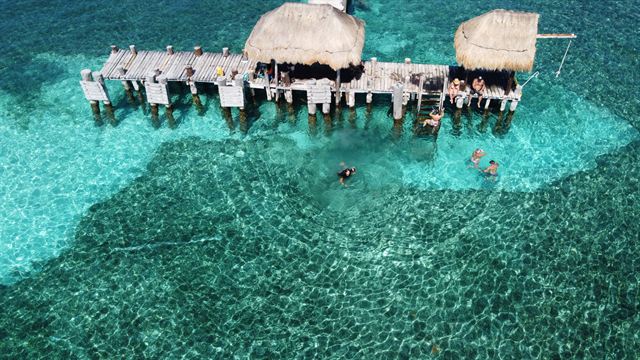 Aerial view of a pier with thatched palapa shelters over clear turquoise shallows at Isla Mujeres