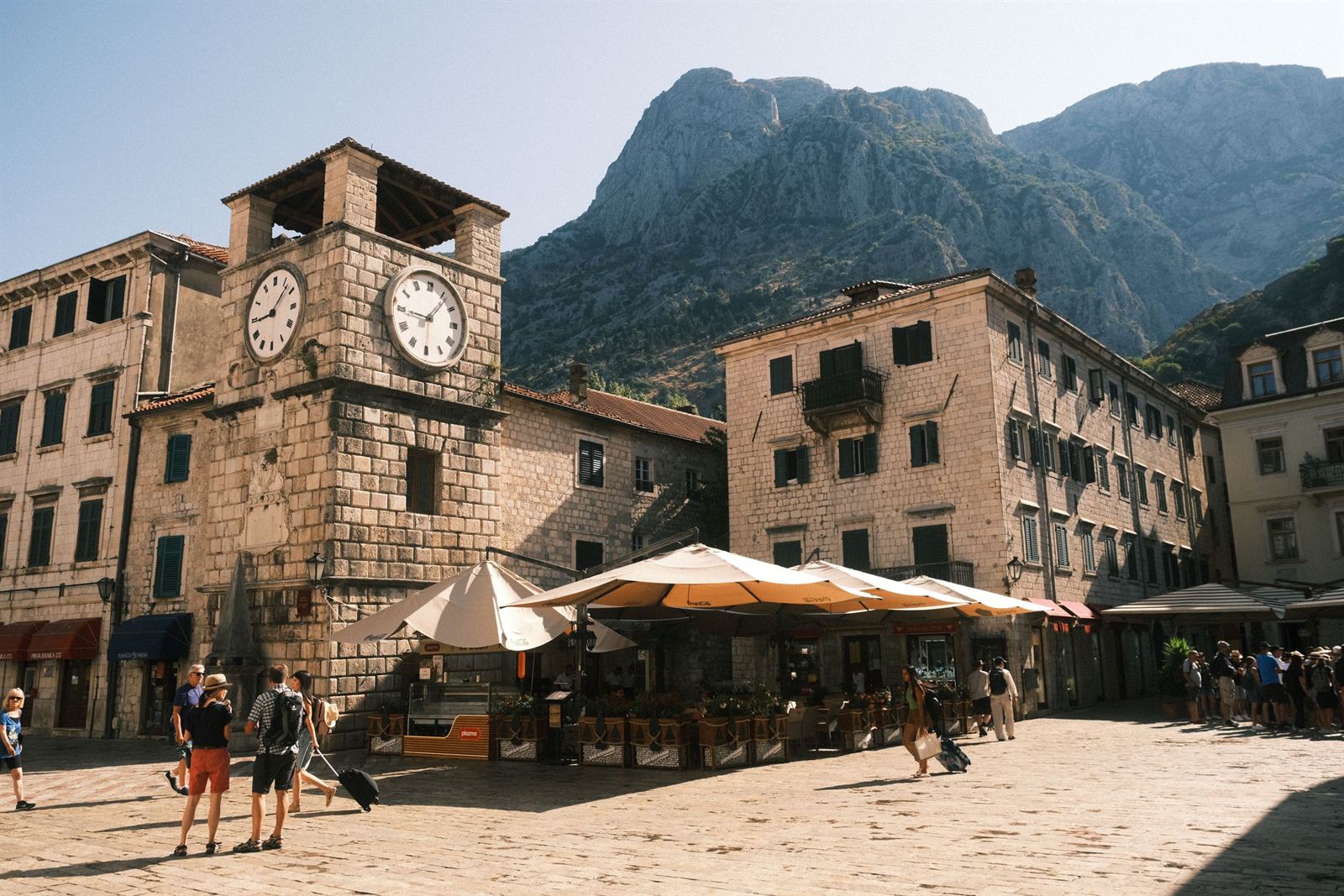 Kotor Old Town Square of Arms with the stone clock tower, cafe umbrellas and mountains behind