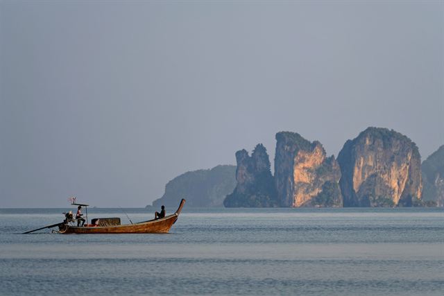 Longtail boat crossing calm water toward jagged limestone island cliffs off Krabi under grey sky