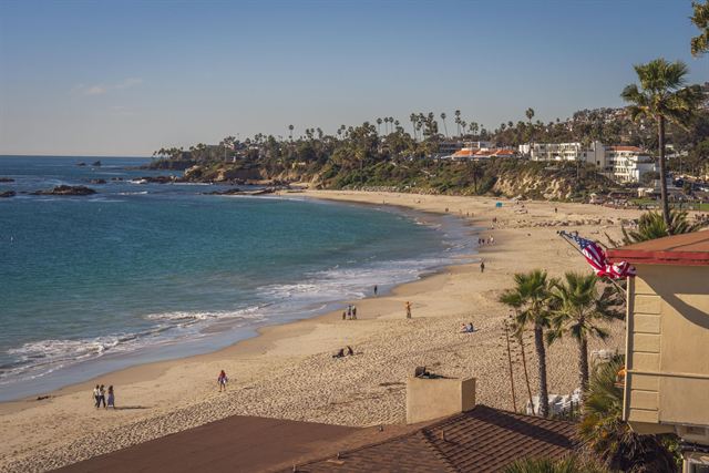 Curving sandy Main Beach area of Laguna Beach with palm trees and hillside homes on a sunny day