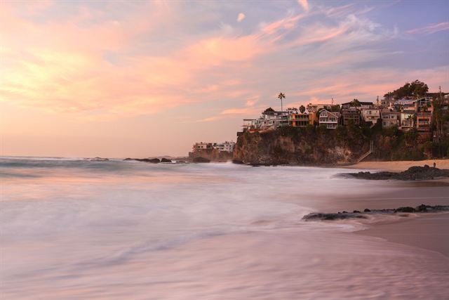 Laguna Beach cliffs topped with homes silhouetted against a pink and orange sunset over wet sand