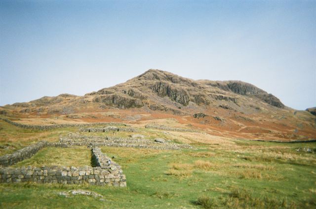 Stone walls of Hardknott Roman Fort on a grassy hillside with a Lake District fell behind