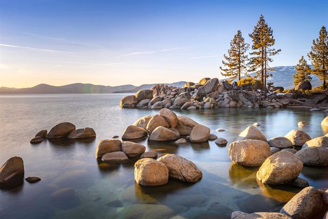 Granite boulders scattered in clear shallow water along Lake Tahoe's shore under pines at sunset