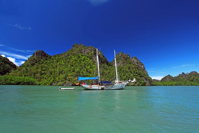 Sailboat anchored in green water below forested limestone hills of Langkawi under blue sky