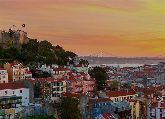 Old Lisbon rooftops below Sao Jorge Castle at sunset with the 25 de Abril Bridge in distance