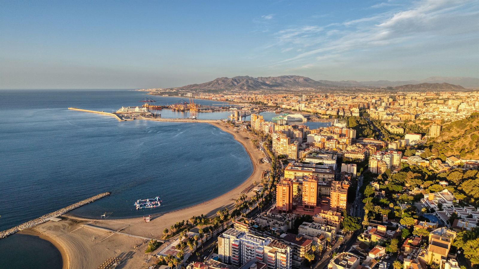 Aerial view of Málaga's curving Malagueta beach, port and city skyline at golden hour