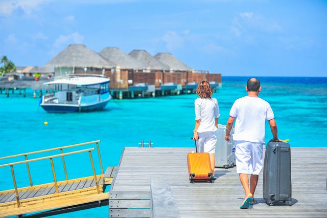 Couple wheeling suitcases along a jetty toward overwater villas and a boat in the Maldives