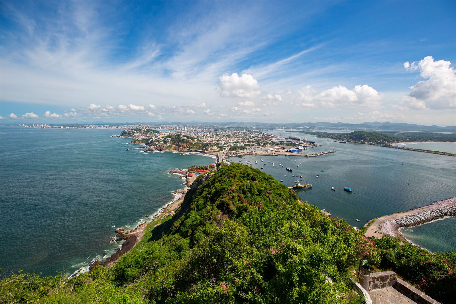Aerial view from a green hilltop over Mazatlan city, harbor channel and Pacific coastline under a cloud-streaked sky
