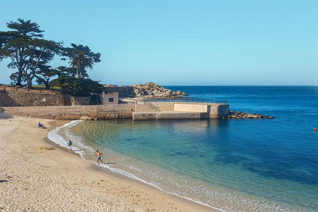 Sandy cove on the Monterey Peninsula with cypress trees, a stone breakwater and clear blue water