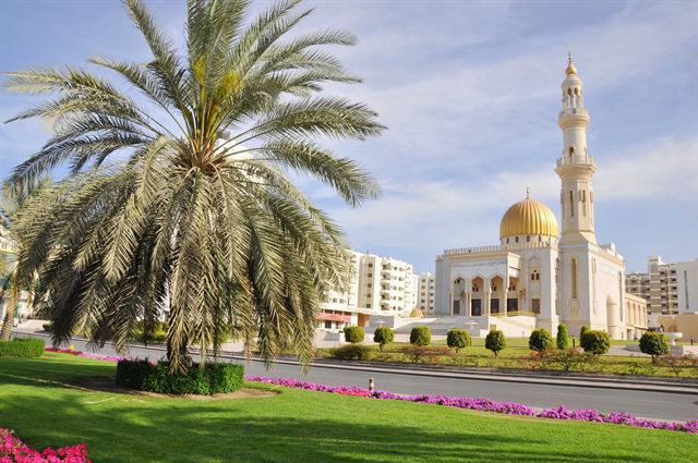 White Zawawi Mosque with its golden dome and minaret behind palm trees in Muscat, Oman