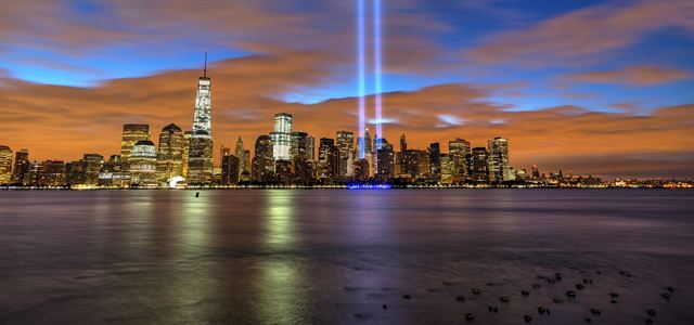 Lower Manhattan skyline across the water at dusk with twin beams of light rising into the sky
