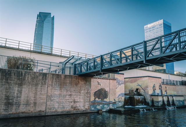 Bricktown Canal in Oklahoma City with a steel footbridge, bison murals and Devon Energy Center tower beyond