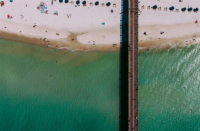 Aerial view straight down on a Panama City Beach fishing pier over turquoise water and sand dotted with umbrellas