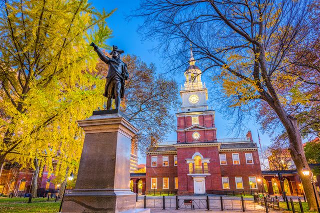 Independence Hall's red-brick clock tower behind a statue, framed by autumn trees in Philadelphia