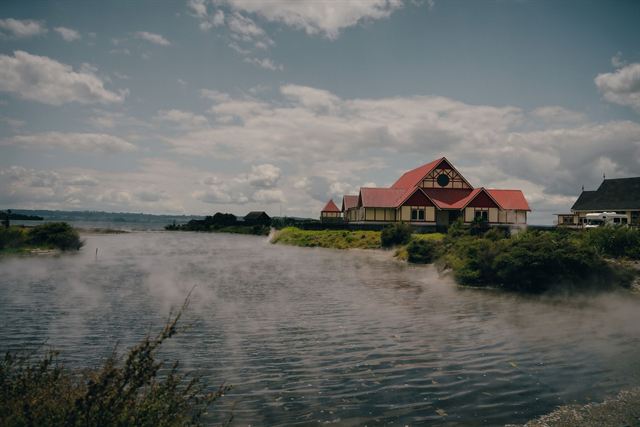 Red-roofed building on the Lake Rotorua shore with geothermal steam drifting over the water