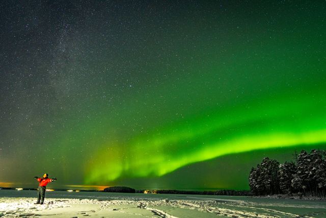 Green northern lights over a snowy Rovaniemi landscape with a lone figure in red