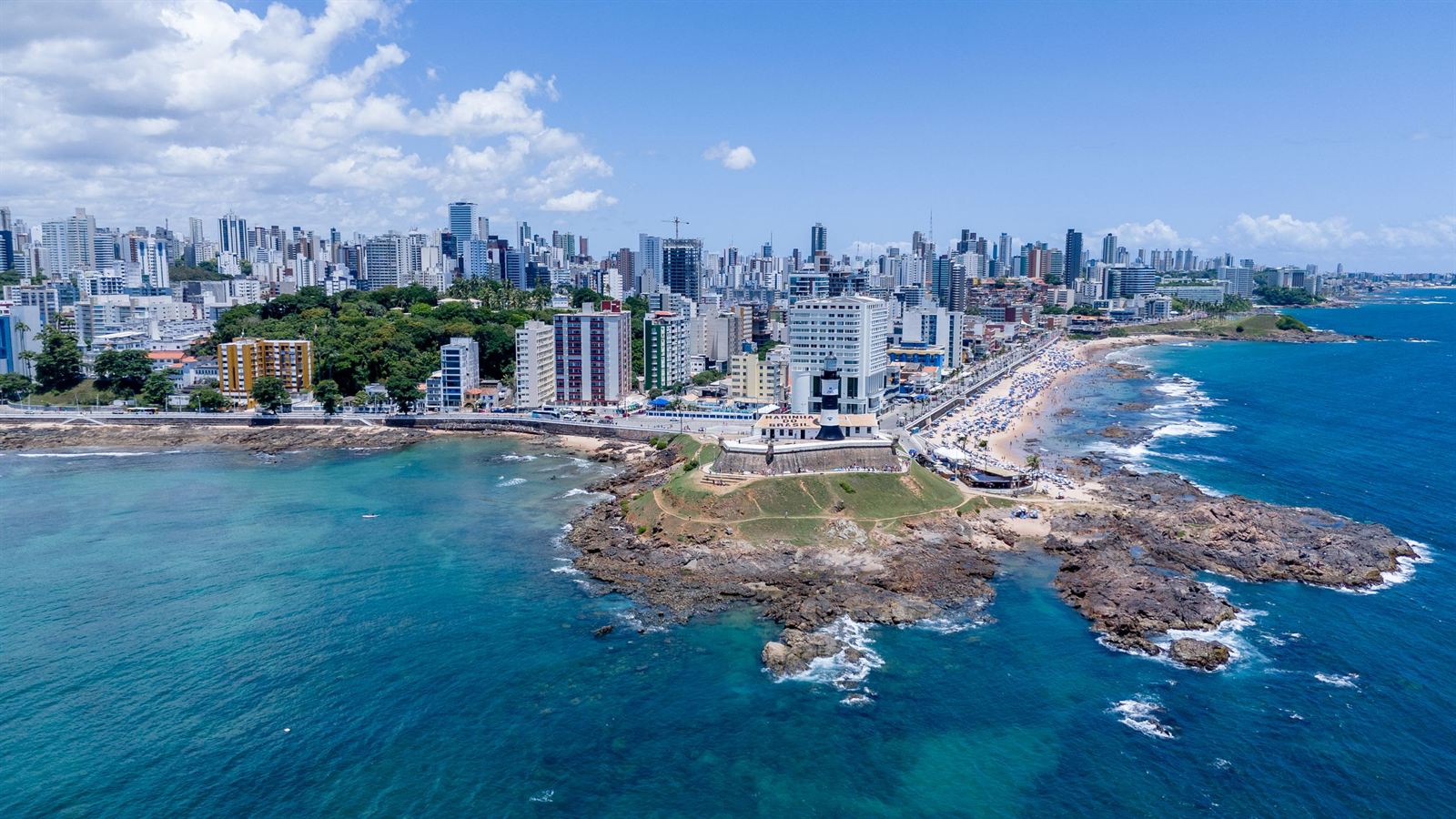 Aerial view of Barra Lighthouse and fort on its rocky point with the Salvador skyline and beaches behind
