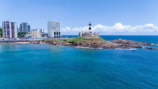 Barra Lighthouse on its green headland beside blue Atlantic water with Salvador high-rises behind