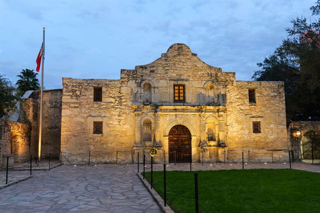 Floodlit limestone facade of the Alamo mission church in San Antonio at dusk beside a Texas flag