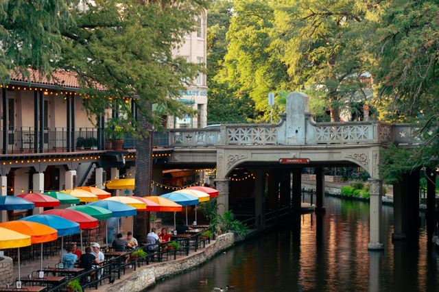 Colorful umbrellas at riverside restaurants beside an arched stone footbridge on the San Antonio River Walk