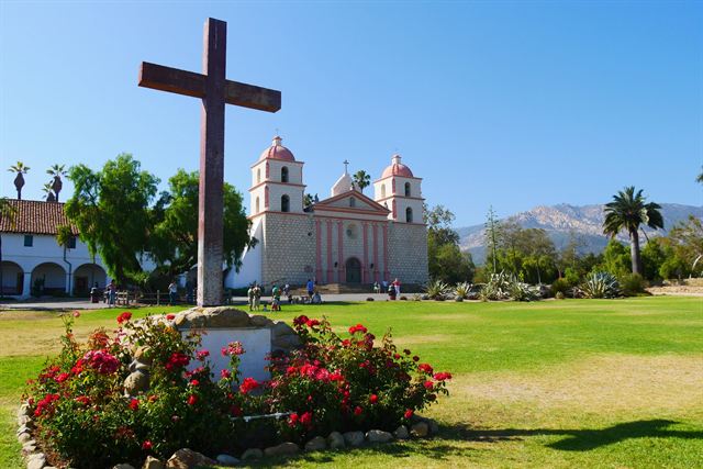 Old Mission Santa Barbara's twin pink bell towers behind a wooden cross, roses and lawn