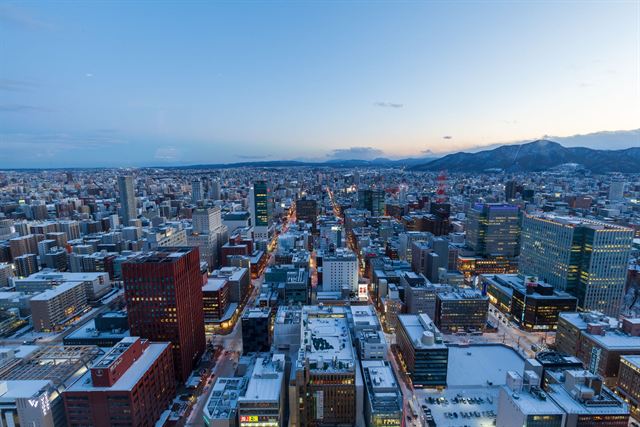 Elevated winter view over snowy Sapporo's grid of city blocks toward mountains at dusk